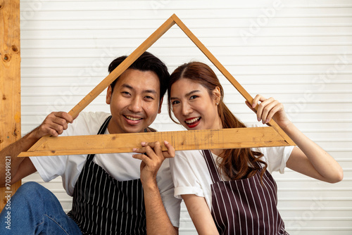 Couple Building Future Together: In Striped Aprons and Jeans Holding a Wooden House Frame Indoors.