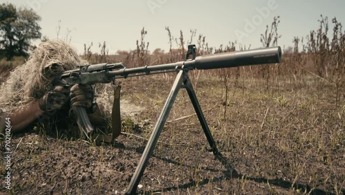 A close-up of a sniper wearing a camouflage suit or grass cloak, waiting and looking through a rifle scope. The sniper aims and shoots. Marksmanship shooting. Cinematic shot photo