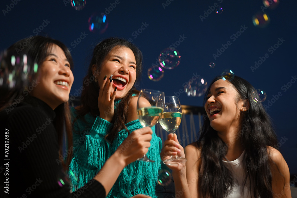 Obraz premium Group of cheerful female friends having fun toasting champagne and laughing together at the rooftop night outdoor party.