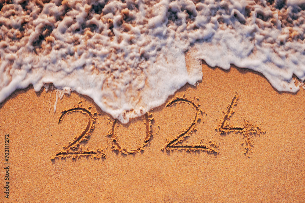 Fototapeta premium New Year 2024 text written on the beach sand and wave at sunrise