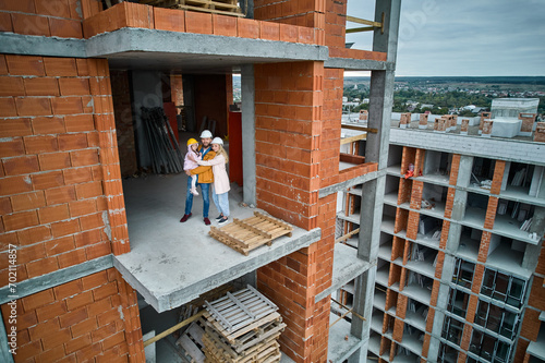 Woman hugging husband and child while standing on concrete floor inside residential building under construction. Family posing in newly constructed apartment building. Drone view