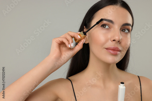 Beautiful young woman applying mascara on grey background