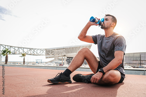 Weary sports man resting and drinking water after endurance workout sitting on the floor. Exhauted male recovering and having a routine break taking a protein supplement at cardio wellness training