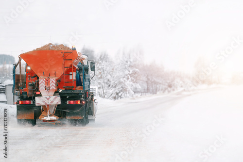 An orange truck with a salt and sand spreader clears the snow and ice from the road. The winter service vehicle works to make the public roads safe and prevent corrosion.