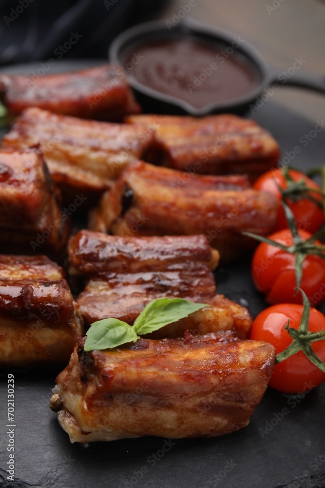 Tasty roasted pork ribs served with sauce, basil and tomatoes on table, closeup