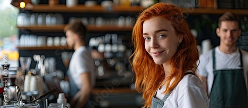 Beautiful Caucasian rad haired lady looks smiling at camera offers hot coffee at modern coffee shop barista posing near counter wearing white t shirt and apron. with copy space image