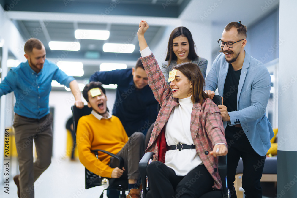 Obraz premium A group of young colleagues are having fun in the office corridor. Young business people ride on office chairs and laugh. Concept of teamwork, relaxation or fun.