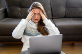 Stressed upset young woman sitting on the floor at home with a laptop and holding her head