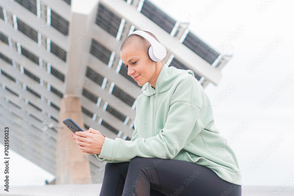 Mid adult woman sitting in stairs after jogging exercise, watching her smartphone and listening music with headphones. Runner female having a break watching content with the phone and smiling