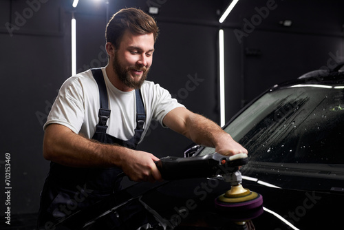Smiling Man in uniform with orbital polisher in repair shop polishing car. Car detailing concept. Portrait of handsome bearded guy at work, applying polisher on car surface. copy space photo