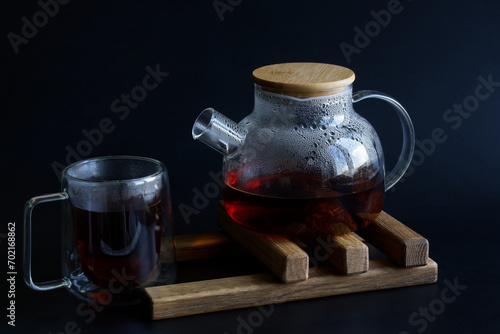 Heat-resistant glass teapot with hot black tea and a heat-resistant mug on a wooden stand for hot dishes. Sweat on the glass walls of the teapot. Black background. Photo. Copy space.