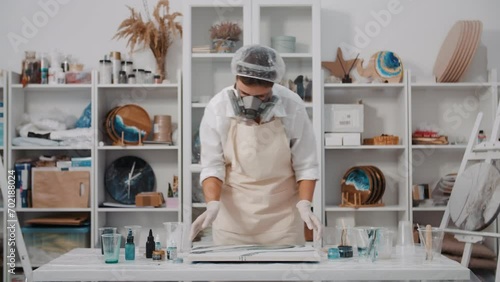 Female artist pours colored art epoxy resin on canvas mixing colors in art studio workshop, creative woman in protective face mask, trendy modern hobby creating beautiful sea waves acrylic paintings photo