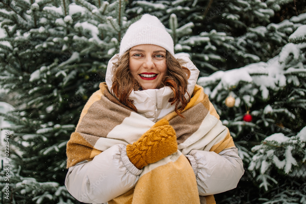 Fototapeta premium Portrait of smiling happy caucasian woman in hat with mittens in winter coat laughing, tossing snow up outside in the city park in winter. High quality photo