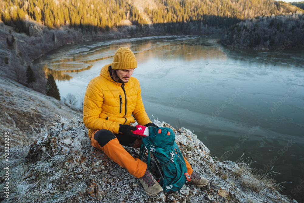 A guy sits in the mountains and takes a first aid kit out of his backpack, a mountain guide provides first aid on a hike, trekking in a mountainous area against the background of a lake. A guy sits in the mountains and takes a first aid kit out of his backpack, a mountain guide provides first aid on a hike, trekking in a mountainous area against the background of a lake.