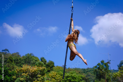 Beautiful Asian girl shows extreme advances by performing pole dance aerial in various positions and spinning stunts on the blue sky around the natural fresh and the waterfall, outdoors.