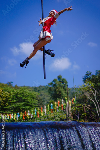 Beautiful Asian girl shows extreme advances by performing pole dance aerial in various positions and spinning stunts on the blue sky around the natural fresh and the waterfall, outdoors. © pomphotothailand