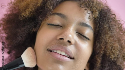 Black woman runs brush over face with eyes closed, smiling enjoying process of applying makeup. African-American happy glad delight girl gets indescribable satisfaction from pleasant touches on skin photo