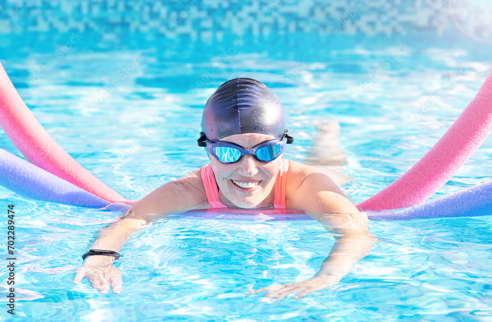 Active exercise senior woman (over age of 50) in goggles, cap doing water sport, aerobics with swim noodles in the swimming pool. Healthy lifestyle.