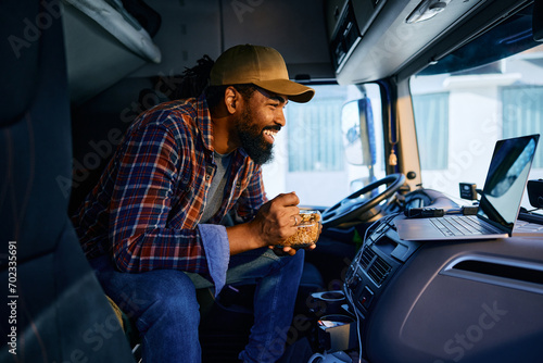 Happy black driver using laptop while eating lunch in his truck cabin. photo