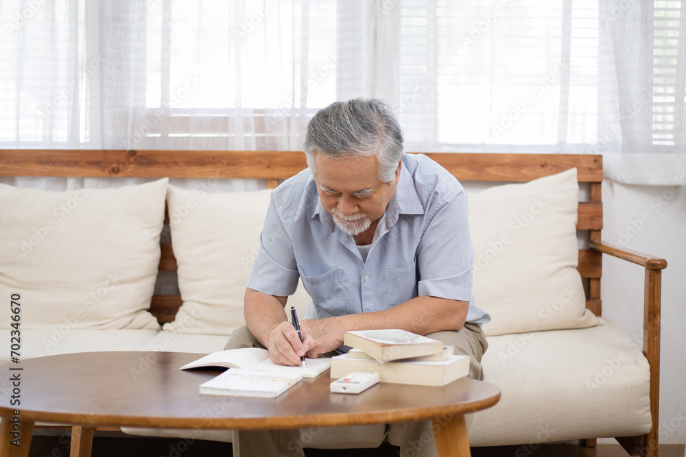 Asian senior man sitting on couch reading, writing or working in living room at home. Doing financial document. retired people living lifestyle insurance concept Asian senior man sitting on couch reading, writing or working in living room at home. Doing financial document. retired people living lifestyle insurance concept