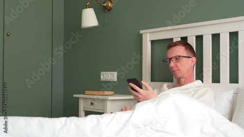 Insomnia. Man with glasses is lying in bed and looking at information on his phone. Interior of modern bedroom. Natural fabrics and materials. Sleep problems. Bleachers for laundry. 4k footage.