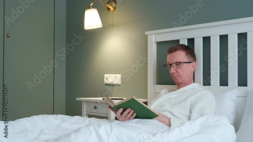 Relaxation and rest. Man with reading glasses is lying in bed and reading book. Interior of modern bedroom. Natural fabrics and materials. Sleep problems. Bleachers for laundry. 4k footage.
