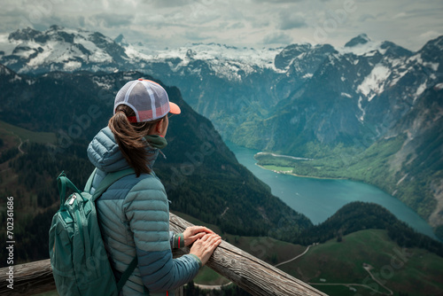 Female hiker with backpack looking at turquoise lake Königssee from above viewpoint of mountain peak Jenner at Berchtesgaden Bavaria, snow-capped mountains in the background. photo