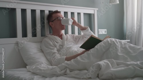 Man is lying in bed, reading book and drinking hot drink, tea, milk, soothing decoction from white ceramic cup. Bedroom interior. Natural fabrics and materials. Sleep problems. 4k footag.