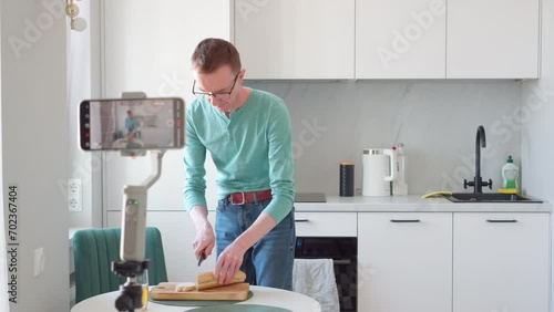 Blogging. Lonely man in turquoise T-shirt and jeans in morning, in kitchen, he films preparation of toast on phone camera. Modern technologies. Professions online. Devices and gadgets. 4k footage.