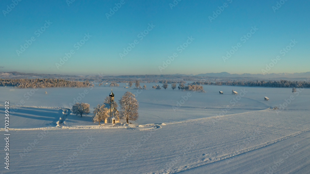 Bavarian church of Raisting with trees and snow during winter and sunset from above, snow field in the foreground, blue sky day, Bavaria Germany.