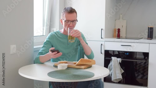 Modern man in long-sleeved T-shirt and jeans is having breakfast in his kitchen, drinking juice and eating toast, looking at phone. Getting information. News online. Rhythm of life. 4k footage.