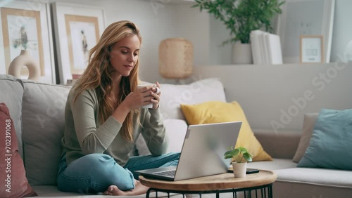 Video of beautiful young woman working with her laptop while drinking a cup of coffee sitting on a couch at home photo