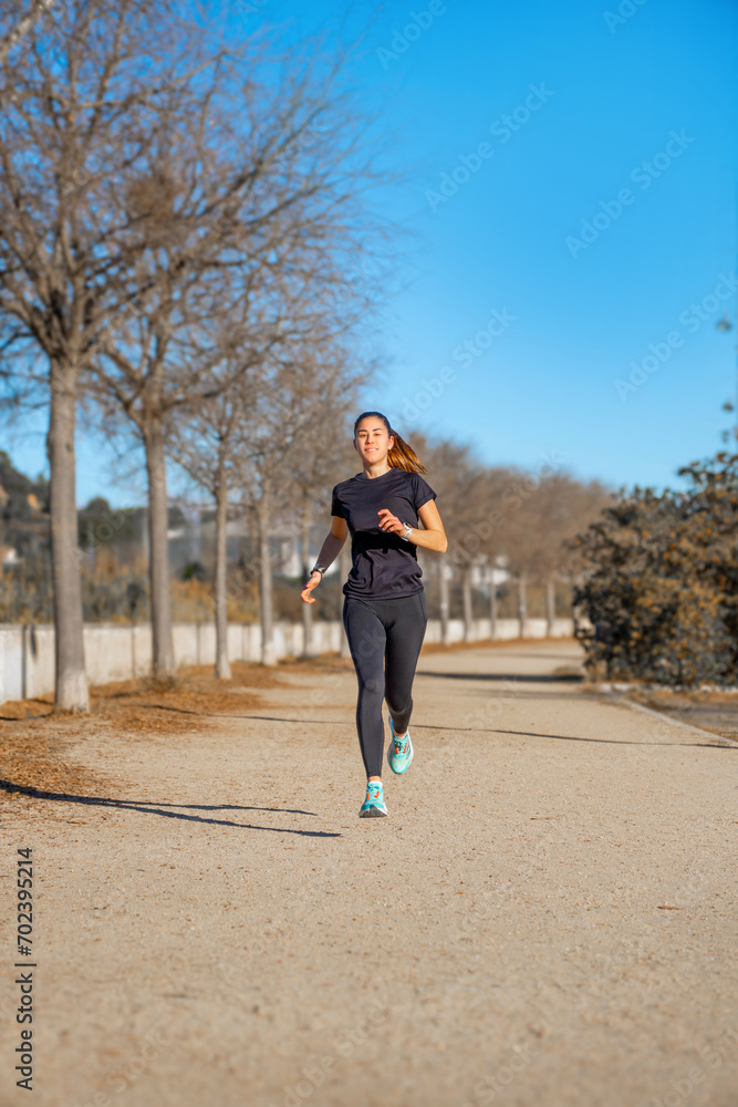 Young female runner, running happy and smiling dressed in black short-sleeved sportswear and tights, suspended in the air training on a dirt walk illuminated by dawn sunlight in a park.
