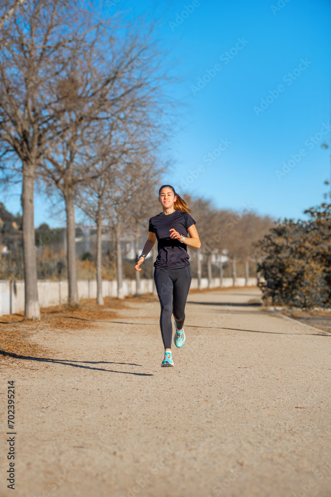 Young female runner, running happy and smiling dressed in black short-sleeved sportswear and tights, suspended in the air training on a dirt walk illuminated by dawn sunlight in a park. Young female runner, running happy and smiling dressed in black short-sleeved sportswear and tights, suspended in the air training on a dirt walk illuminated by dawn sunlight in a park.