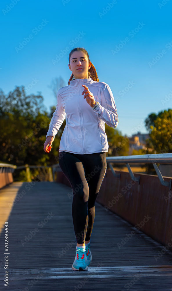 Attractive young runner girl, dressed in black tights and white sports jacket, running training in the morning on a wooden floor bridge under the dawn light of a public park. Attractive young runner girl, dressed in black tights and white sports jacket, running training in the morning on a wooden floor bridge under the dawn light of a public park.