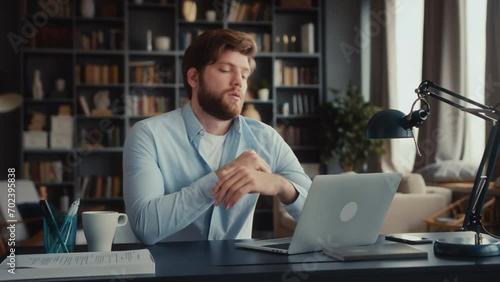 young man with beard in blue shirt sits home office, works at laptop. overworked entrepreneur sleeps with head on desktop. weary and tired worker stretch neck, breath out, starts typing on keyboard photo