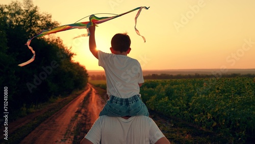 Son plays dreams of fly, traveling, Happy boy on dad shoulders runs along country road, plays with toy kite, sunset. Child, dad is running with kite, sun. Kite floats in child hand. Kid with toy kite