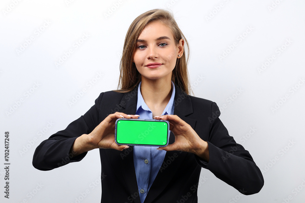 Caucasian fair-haired young woman in a jacket shows a green screen of a smartphone for advertising Caucasian fair-haired young woman in a jacket shows a green screen of a smartphone for advertising
