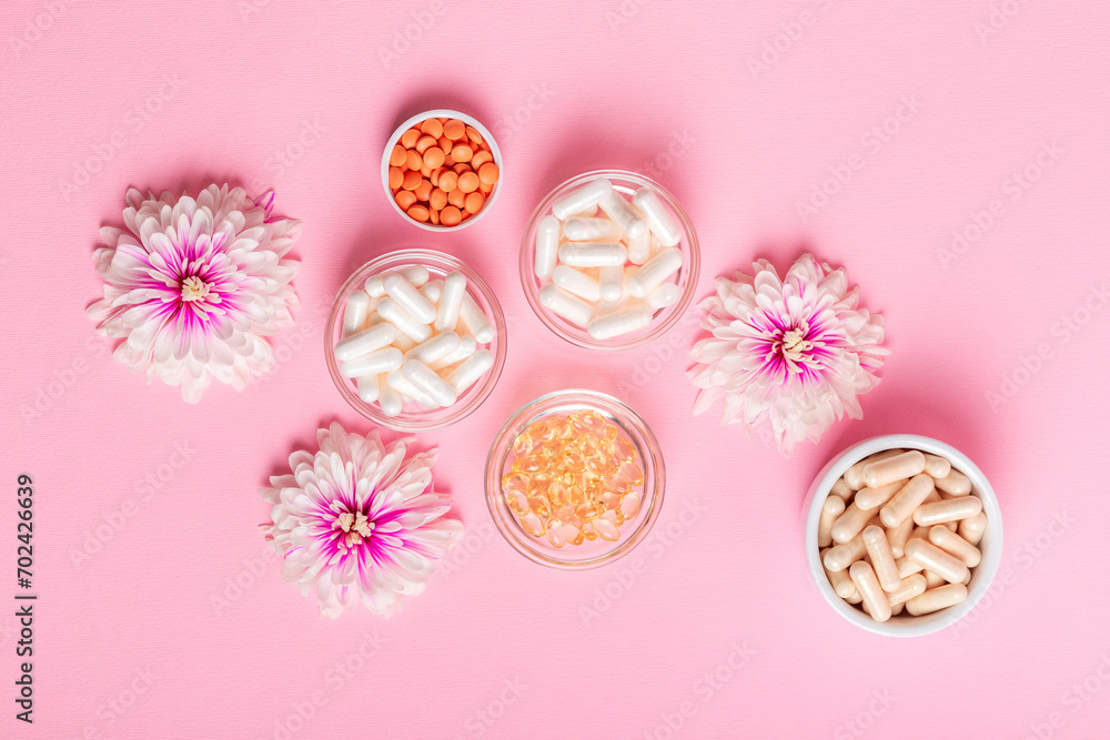 Organic food supplement, minerals and different vitamins such as vitamin D3, C, E in small glass and ceramic jars, pink flowers on pink background top view. Take care of yourself.