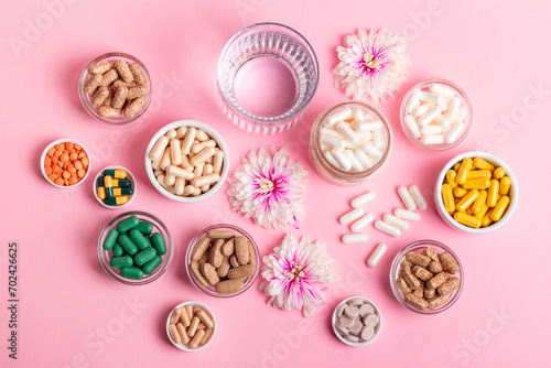 Many vitamins, dietary supplements, pills, meds, capsules and tablets in small jars, pink flowers and a glass of water on pink background top view. Horizontal photo.