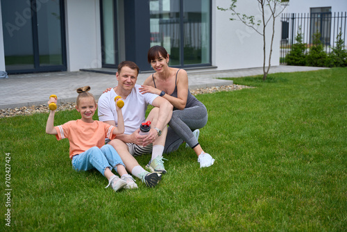 Happy mother, father and daughter sitting on grass and looking at camera in yard