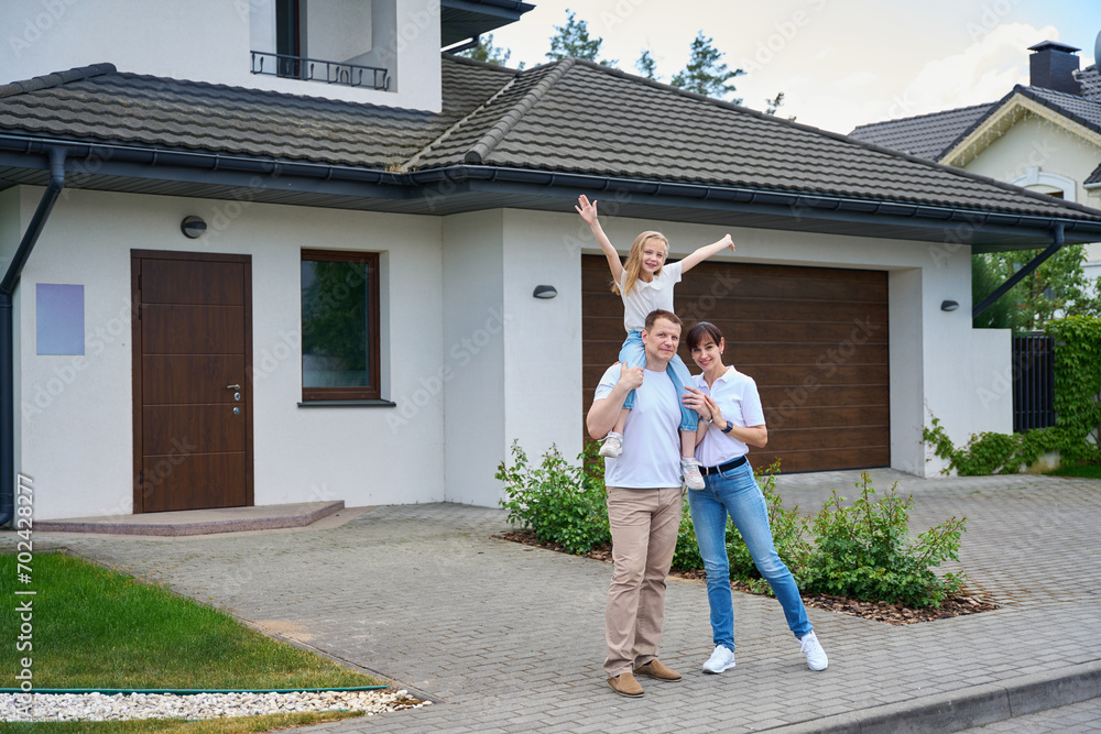 Fototapeta premium Pleased mother and daughter on father's shoulders in front of new townhouse