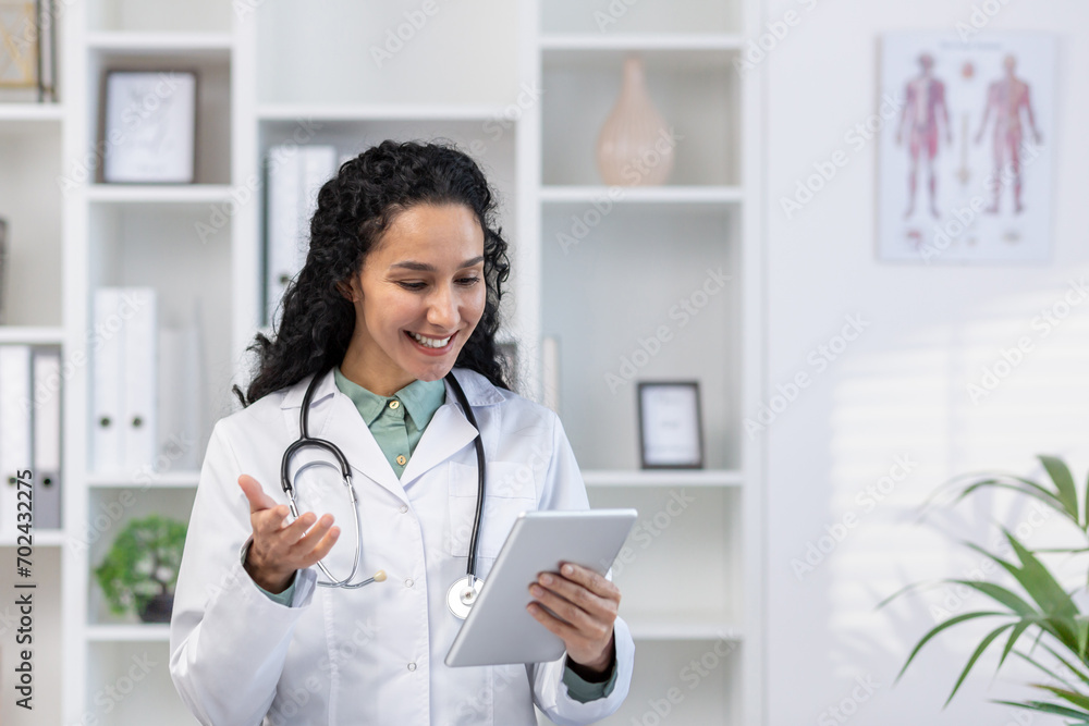 Doctor inside the medical office stands with a tablet computer in hands, a clinic worker in white medical robe will advises the patient remotely using the video for video call, joyfully and friendly. Doctor inside the medical office stands with a tablet computer in hands, a clinic worker in white medical robe will advises the patient remotely using the video for video call, joyfully and friendly.