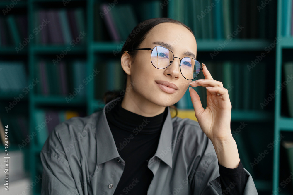 Intelligent young woman adjusting glasses in a library, symbolizing knowledge and study.