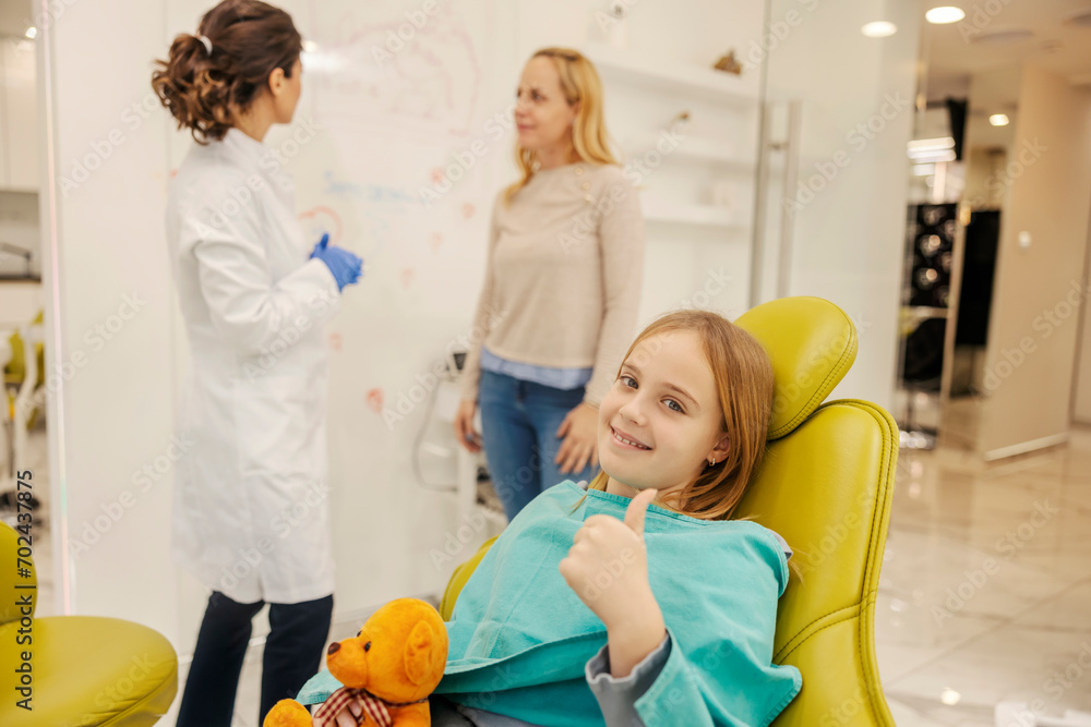 A happy girl giving thumbs up and smiling at the camera at dentist office while mother and dentist talking. A happy girl giving thumbs up and smiling at the camera at dentist office while mother and dentist talking.