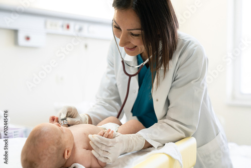 Smiling female pediatrician using stethoscope and protective gloves to check up a newborn baby. photo