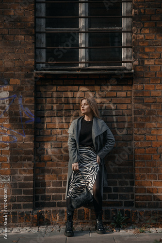 Stylish blond woman posing near red brick wall. Beautiful fashionable girl stands near the brick wall. Fashionable woman in spectacles hanging out in night city.