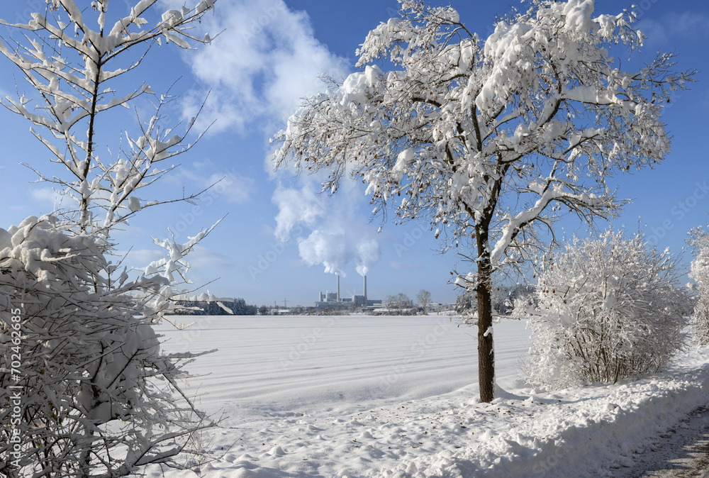 Combined heat and power station at the horizon emitting white smoke on a winter day. Snow covered trees and bushes in the foreground.
