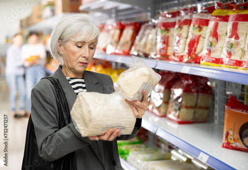 Interested positive senior lady reading label on packages of Asian noodles while shopping in grocery section of supermarket .. photo