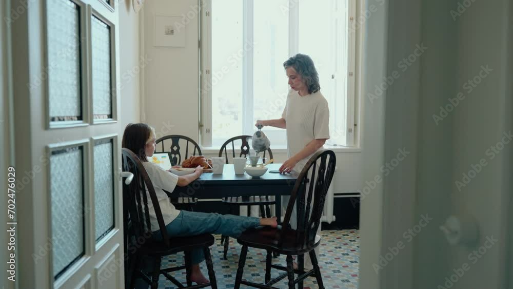 Daughter looking at tablet before eating breakfast with her confident young mom in kitchen Daughter looking at tablet before eating breakfast with her confident young mom in kitchen